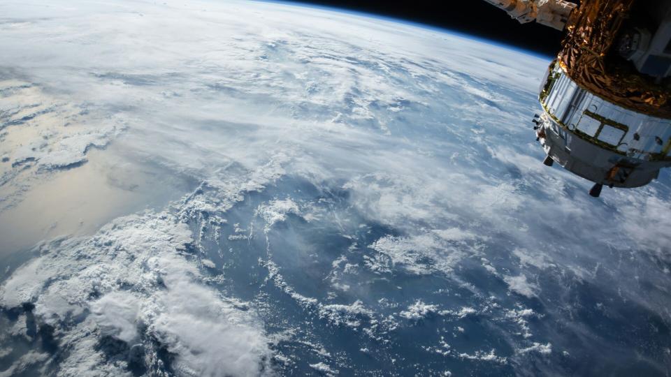 Earth seen from orbit, cloud-covered ocean curving away to a thin blue horizon, with a satellite in the upper right.